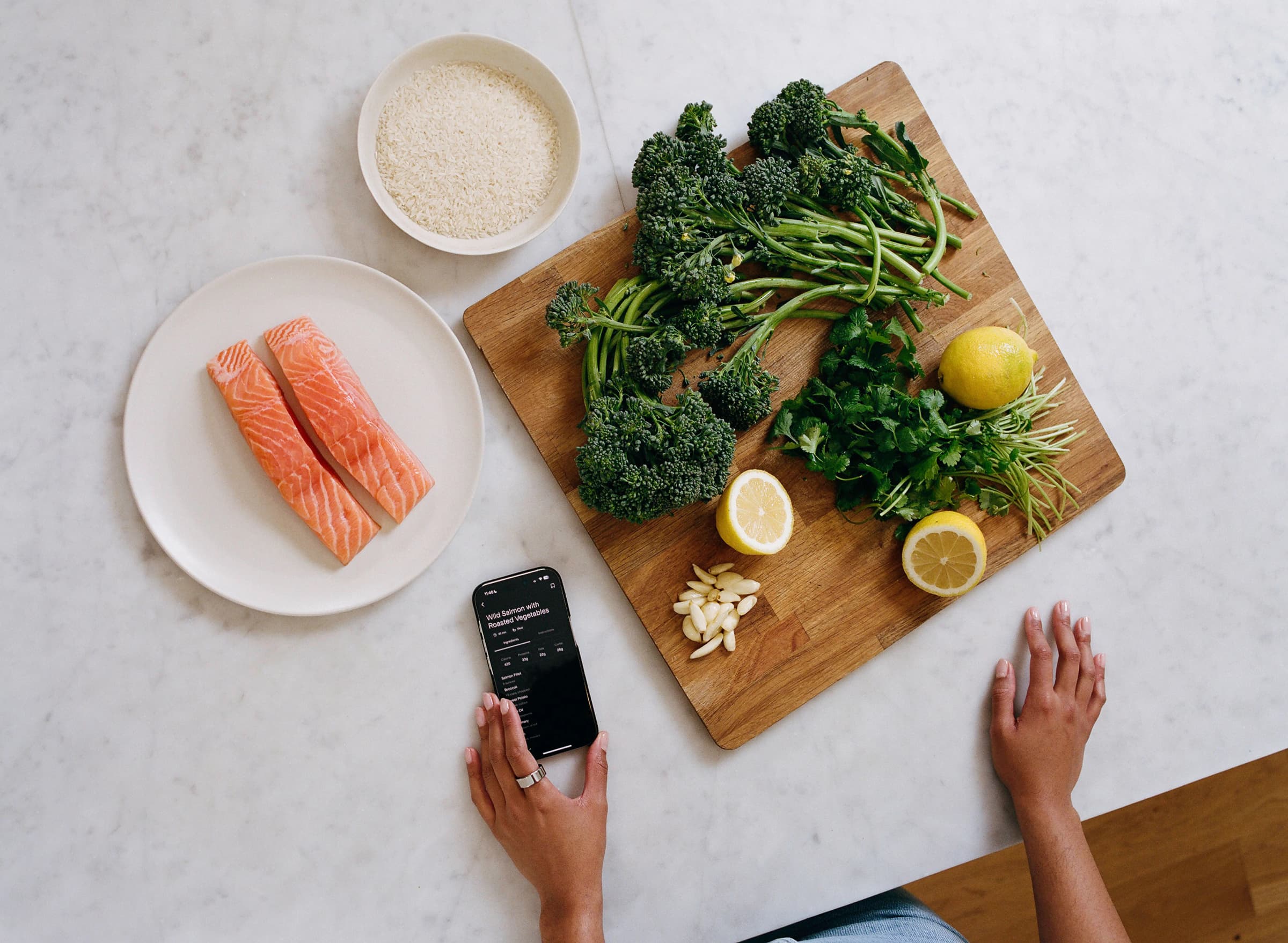 White stone countertop with various vegetables on a cutting board, a bowl of uncooked rice, and a plate of raw salmon