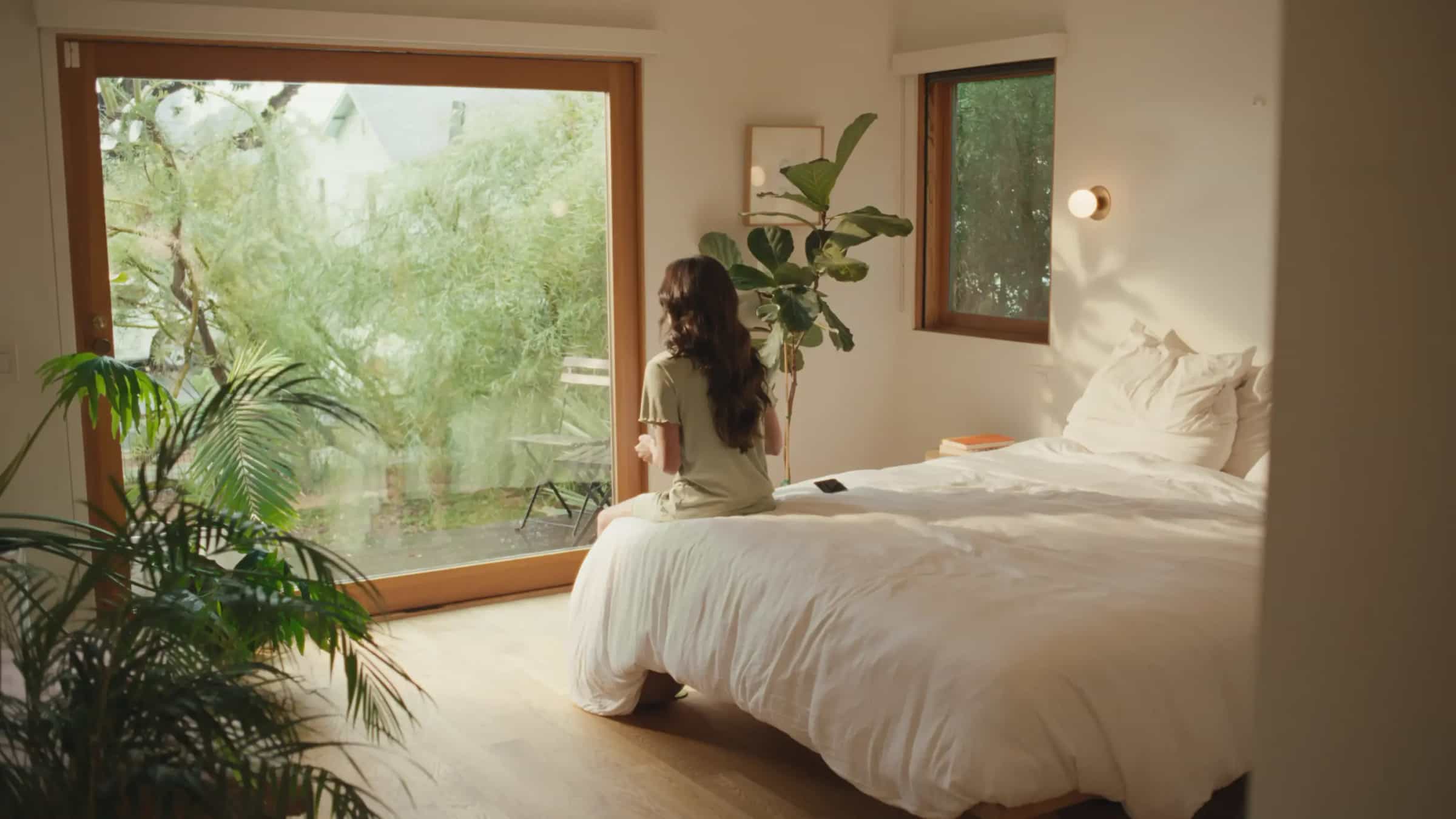 Sunny bedroom with green plants surrounding. Woman sitting on bed starting out large window with trees